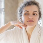 Front view of a woman in her bathrobe, looking in a mirror at her SPMU treatment aftercare.