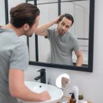 A man standing near the mirror is closely examining his eyebrows. 